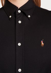 Czarna koszulka polo z teksturowanej tkaniny, klasycznym kołnierzykiem i trzema białymi guzikami. Na piersi ma małe haftowane logo polo z zawodnikiem.