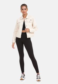 Lichtbeige denimjack met een klassieke kraag, voorzakken en knoopsluiting, gecombineerd met zwarte aansluitende legging en zwarte sneakers.