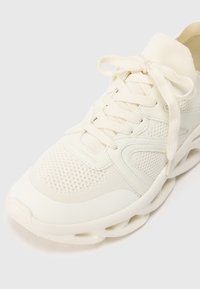 Basket blanc de sport avec tige en maille et tissu, semelle épaisse avec des découpes, et lacets à plat noués sur un fond neutre.