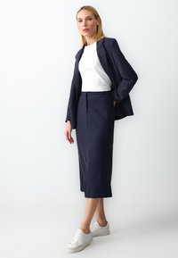 Marineblauwe streepjasje gedragen over een wit topje, gecombineerd met een bijpassende marineblauwe kokerrok en witte sneakers. Glad stoftextuur, nauwsluitende pasvorm.