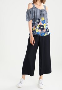 Kwiatowa bluzka na ramionach w kolorach niebieskim, różowym i żółtym z falbanianymi rękawami i wzorzystym deseniem, w połączeniu z czarnymi spodniami szerokimi i białymi trampkami.