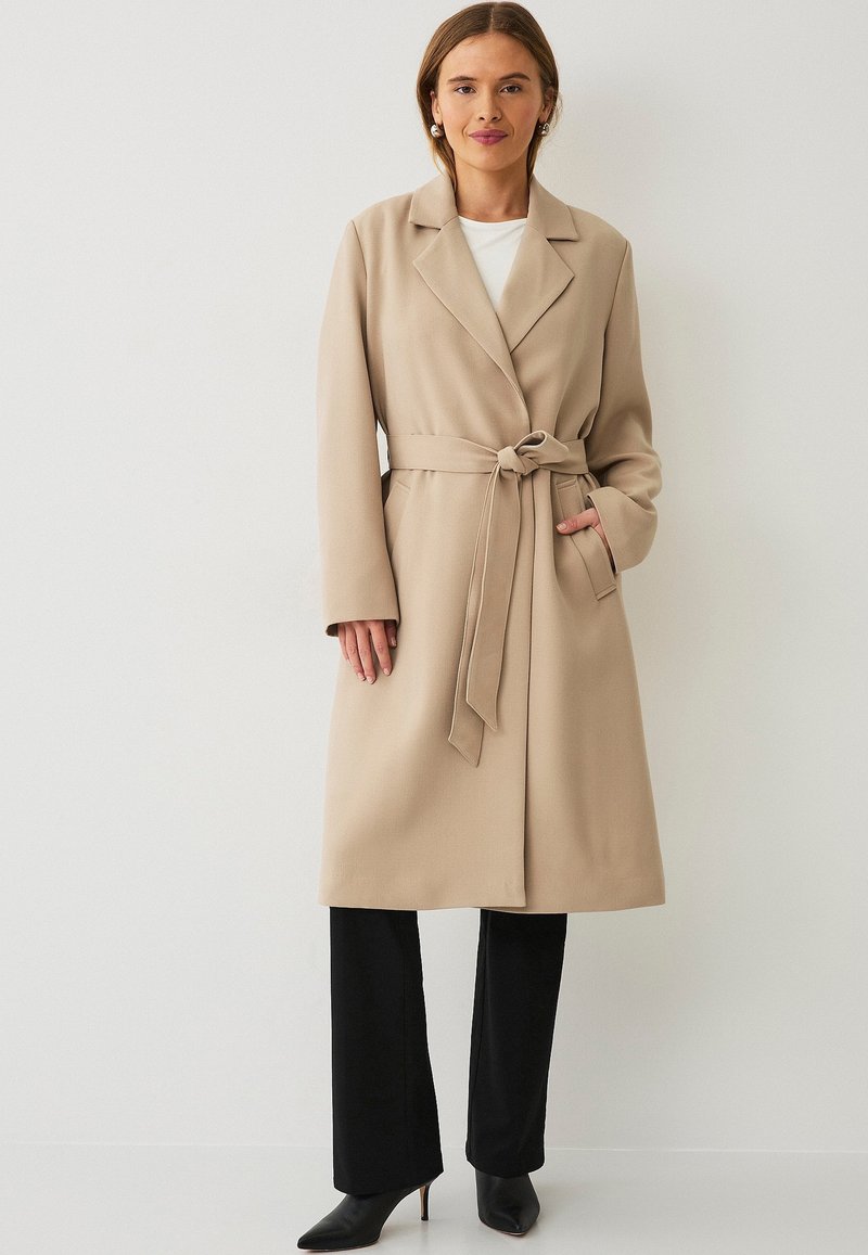 Beige lange jas met een wikkelontwerp, ceintuur in de taille en kraag. Draagt over een wit shirt, gecombineerd met zwarte broek en puntige schoenen.