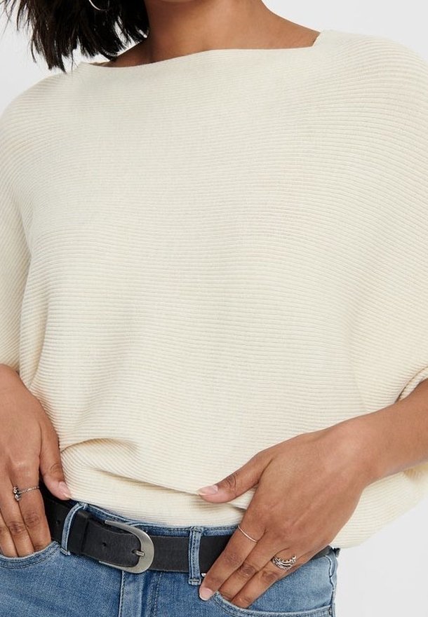 Femme portant un pull côtelé écru rentré dans un jean bleu avec une ceinture noire, montrant des mains ornées de bagues en argent près de la taille.