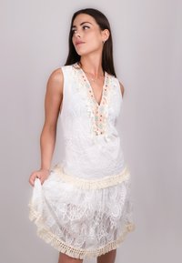 Abito bianco in pizzo con scollatura a V, dettagli floreali e orlo frangiato. Presenta motivi intricati e un design a strati e tessurizzato.