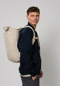 Sac à dos avec un design à rabat, en matériau texturé beige, et des bretelles ajustables. Le modèle porte une veste noire et un pantalon gris, se tenant de profil.