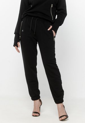 Kobieta ubrana w czarne spodnie dresowe z zamkami błyskawicznymi w kieszeniach oraz dopasowaną czarną bluzę na zamek, uzupełnione czarnymi sandałami na wysokim obcasie.
