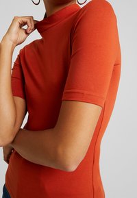 Dopasowana, krótka bluzka w kolorze rdzy, z wysokim dekoltem i gładką fakturą. Ścisłe mankiety podkreślają rękawy.