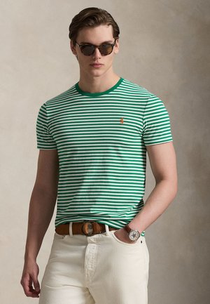 Noor mees rohelise-valge triibulise t-särgi, valgete pükste, pruuni vöö, käekella ja tumedate päikeseprillidega, seismas neutraalse tausta ees.