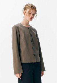 Femme aux cheveux clairs portant une veste brune à boutons et à manches longues, ainsi que des pantalons noirs, se tenant devant un fond blanc uni.