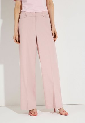 Person trägt weite, hoch taillierte Pastellrosa Hosen mit einem in das Hosenbein gesteckten, hellrosa Hemd und passenden rosa offenen Zehentitzen.