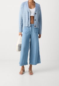 Žena v světle modrém kardiganu, bílém crop topu, širokých džínech, béžových sandálech na podpatku, držící bílou kabelku na jednobarevném pozadí.