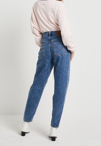 Högmidjade blå denimjeans med avslappnad passform, fem fickor och synliga sömdetaljer. Bärs med en ljusrosa långärmad topp.