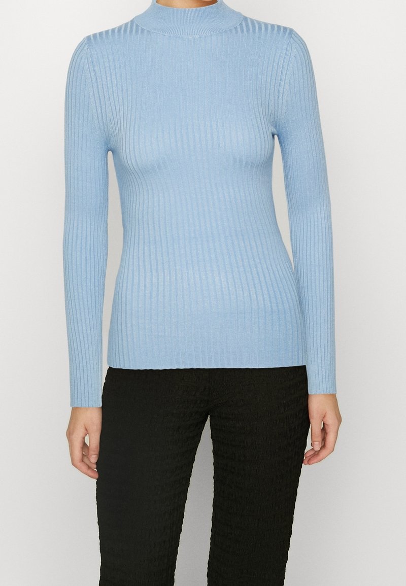 Femme portant un pull ajusté à manches longues côtelé bleu clair et un pantalon noir texturé, debout devant un fond clair uni.