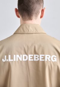 Bežna jakna s ovratnikom in gladko teksturo, ki na hrbtu vsebuje velike bele črke "J.LINDEBERG". Minimalističen dizajn.