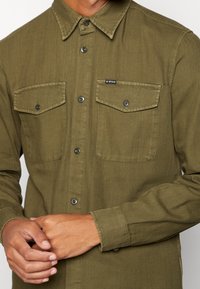 Olivinvihreä, tekstuuri-kangasinen nappipaita, jossa on kaksi rintataskua, musta logolappu ja metalliset napit.