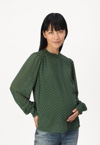 Blouse verte texturée avec un motif géométrique, manches longues, poignets ajustés et col rond. Associée à un jean bleu clair.