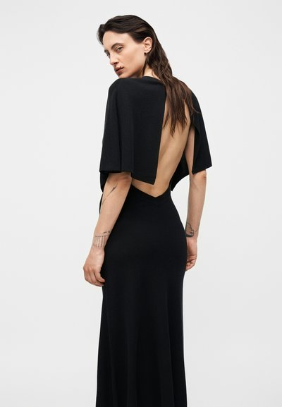 Γυναίκα με μακριά σκούρα μαλλιά που φοράει μαύρο φόρεμα χωρίς πλάτη, γυρισμένη ελαφρώς για να δείξει τατουάζ στα χέρια της με φόντο απλό λευκό.