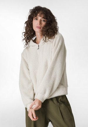Fuzzy cream-colored zip-up hoodie with a lined hood, ribbed cuffs, and textured fabric, paired with olive green pants.