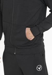 Hoodie zippé noir avec une texture lisse et des poches avant, associé à un pantalon assorti arborant un logo blanc sur la jambe inférieure.
