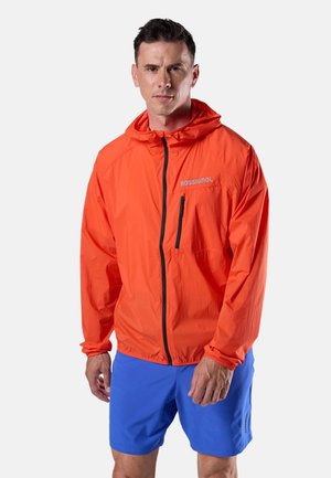 Man die een feloranje capuchonjas met zwarte rits en blauwe korte broek draagt, staand tegen een effen lichte achtergrond.