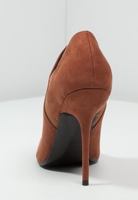 Scarpa con tacco alto in suede marrone, con punta affilata, superficie liscia e tacco slim nero. Presenta una striscia decorativa orizzontale marrone.