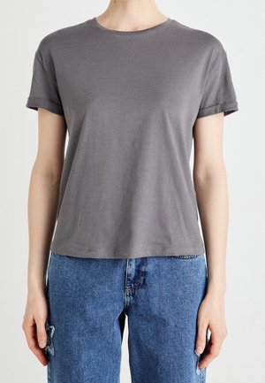 Osoba ubrana w gładką szarą koszulkę z krótkim rękawem i niebieskie dżinsy z kwiatowymi naszywkami po bokach, stojąca na białym tle.