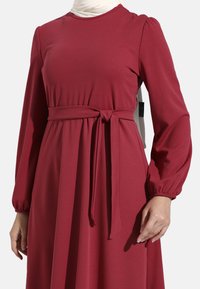 Langdurige bordeauxrode jurk van gestructureerde stof, met een gestrikte taille en pofmouwen, een ronde halslijn en een vloeiende pasvorm.