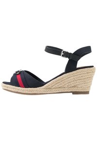 Sanda na klínu s námořnickými látkovými popruhy, červeným akcentem a tkanou juteovou podrážkou. Popruh kolem kotníku s nastavitelnou přezkou a otevřenou špičkou.