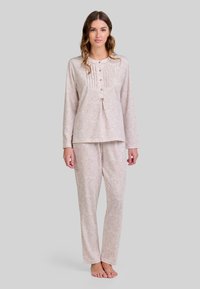 Mujer de pie descalza, vistiendo una camisa de pijama de manga larga y botones en color beige claro, y pantalones de pijama largos a juego con un pequeño patrón de puntos.