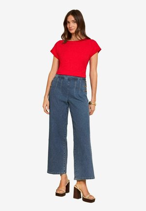 Žena nosiaca červený krátky rukáv, modré široké rifle s vysokým pásom a bočnými gombíkmi, béžové platformové lodičky a zlatý náramok.