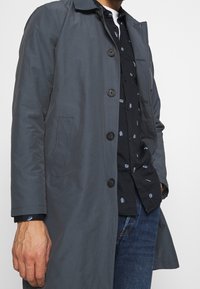 Cappotto grigio realizzato in tessuto liscio, con chiusura a bottone, tasche laterali e colletto classico, indossato sopra una camicia nera con motivo.