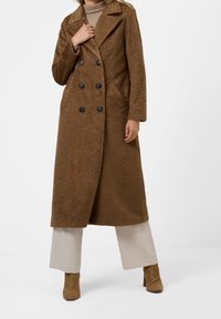 Brun lång kappa i strukturerad tyg, med dubbelknappling, svarta knappar och sidofickor, kombinerad med ljusa byxor och klackboots.