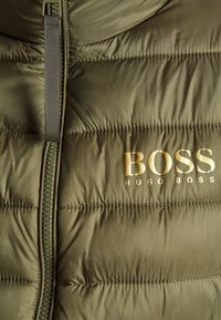 Sección de chaqueta acolchada en verde oliva con cremallera frontal y logo dorado "BOSS HUGO BOSS" en el lado derecho.