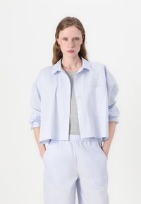 Camisa azul clara com riscas, de corte descontraído, com bolso à frente e mangas enroladas, combinada com uma blusa cinza de mangas curtas e calças a combinar.