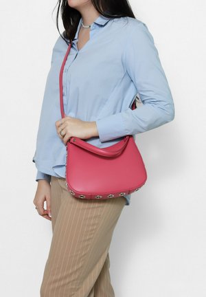 Vrouw die een lichtblauw overhemd draagt en beige broek met krijtstreep, met een roze schoudertas met metalen oogjes aan de onderkant.