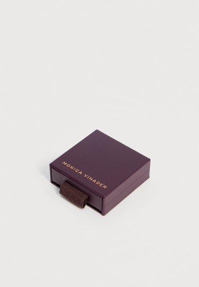 Μικρό σκούρο μωβ τετράγωνο κουτί κοσμημάτων με υφασμάτινη λαβή και το όνομα "Monica Vinader" τυπωμένο με χρυσά γράμματα στο καπάκι.