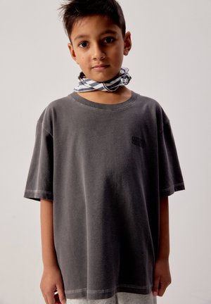 Ragazzo con capelli corti e scuri indossa una maglietta grigio scuro oversize e una sciarpa a quadri bianca e nera legata intorno al collo, in piedi davanti a uno sfondo semplice.