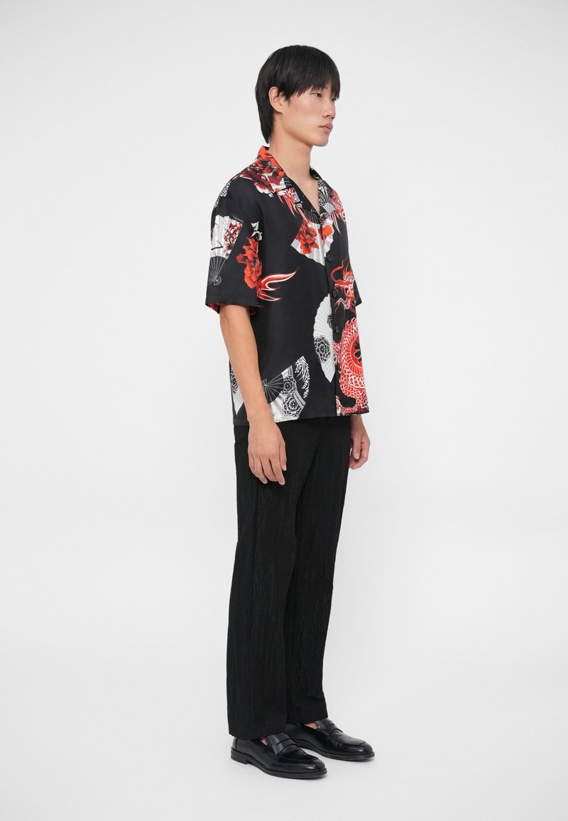 Kurzärmliges schwarzes Shirt mit roten und weißen Blumen- und Fächermustern, kombiniert mit strukturierten schwarzen Hosen und schwarzen Slippern.