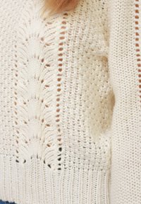 Strikket sweater i cremefarve med åbne strikkemønstre og ribstrikkede manchetter. Blød tekstur med subtile variationer i strikkedesignet.