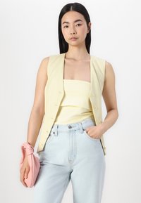 Un gilet sans manches jaune clair par-dessus un top assorti, associé à un jean taille haute bleu clair. Le modèle tient une pochette rose.