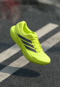 Helles gelbes Sportschuh mit schwarzen Streifen, leichtem Mesh-Obermaterial, abgerundeter Zehenpartie und strukturierter Sohle, für hohe Leistung konzipiert.