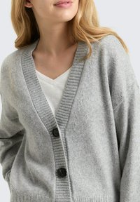 Cardigan gris clair en tissu tricoté doux, avec un col en V profond, des poignets côtelés texturés et deux boutons foncés à l'avant.