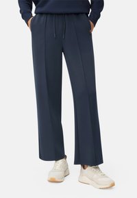 Marineblauwe wijde broek met een trekkoord in de taille, zijzakken en een gladde textuur, gecombineerd met crèmekleurige sneakers en een donker sweatshirt.