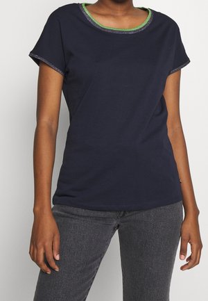 Granatowa koszula z krótkim rękawem z zielonym i srebrnym obszyciem na kołnierzu i rękawach, noszona z ciemnoszarymi dżinsami.