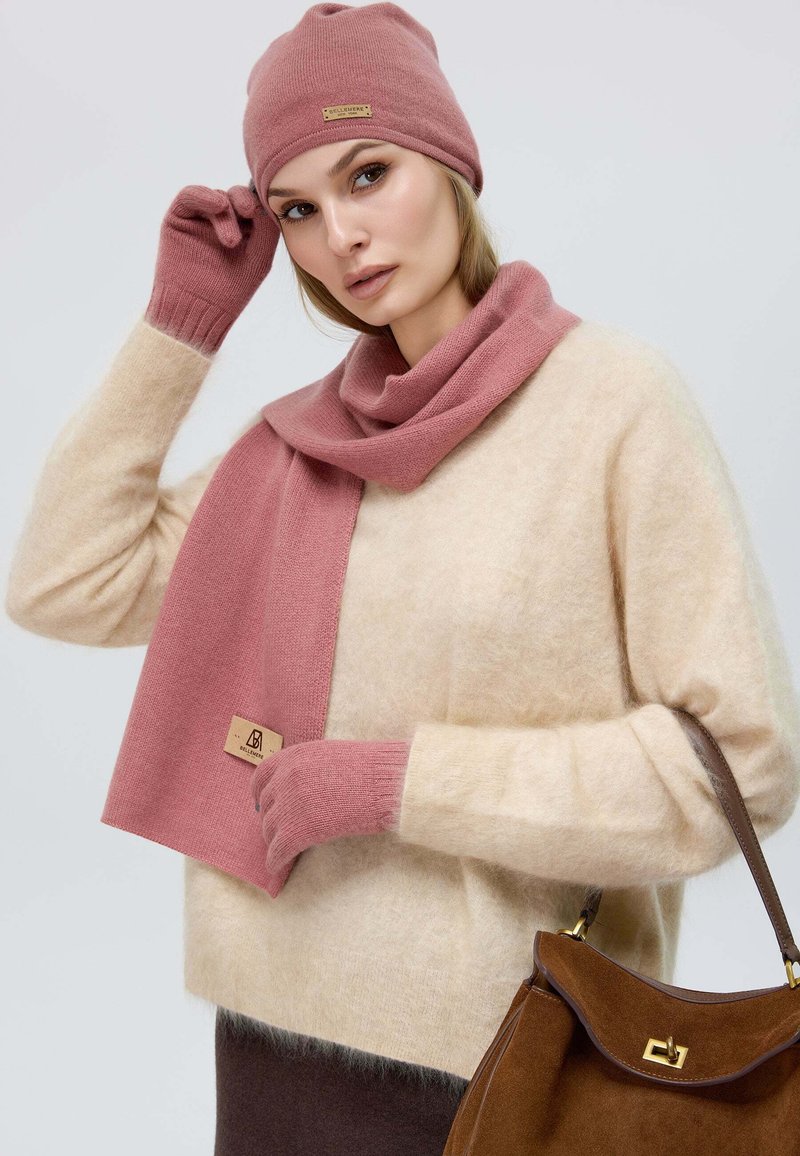 Rosa Strickmütze, passende Schal und Handschuhe; weiche Textur. Cremefarbener Pullover mit rosa Akzenten, braune Handtasche, neutraler Hintergrund.