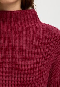 Gros plan d'une personne portant un pull en maille bordeaux à col haut, montrant le motif côtelé texturé et la zone des épaules.