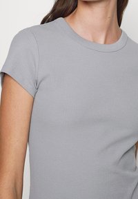 Kobieta nosząca jasnoszary, krótki rękaw ribbed t-shirt z okrągłym dekoltem, pokazana od ramienia do połowy torsu.