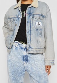 Vaalean sininen farkkutakki, jossa on sherpa-kaulus, kaksi rintataskua ja etunapit. Yhdistetty mustan paidan ja vaaleansinisien farkkujen kanssa.