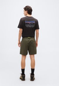 Άνδρας που φοράει μαύρο μπλουζάκι Patagonia με λογότυπο βουνού και ελιά πράσινο σορτς, στέκεται με την πλάτη στραμμένη σε ένα απλό φωτεινό φόντο.