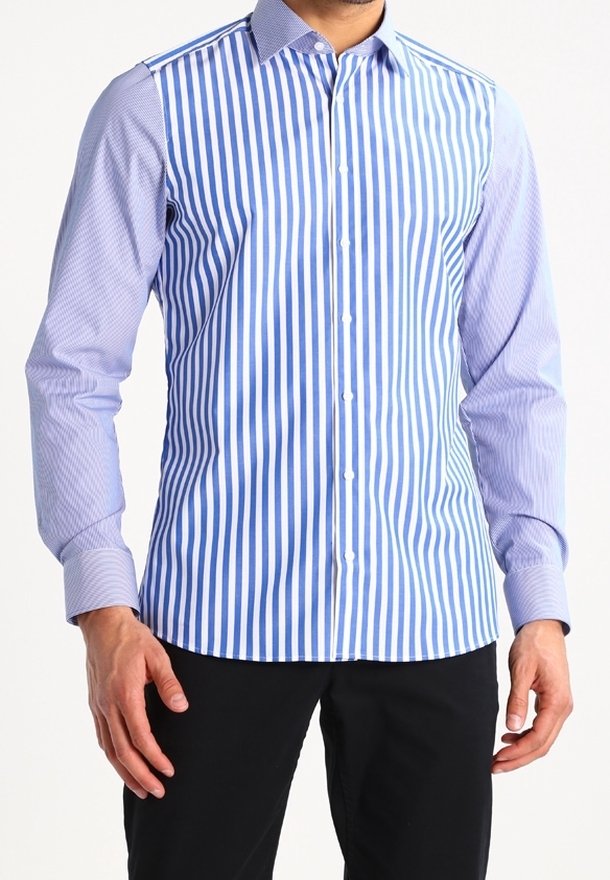 Homme portant une chemise boutonnée avec des rayures verticales bleues et blanches sur le devant et des manches longues bleu clair à motifs, associée à un pantalon noir.